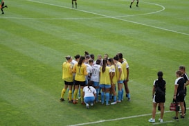 A group of female soccer players, wearing yellow and white jerseys, stands in a tight huddle on a lush green soccer field. The players are focused and engaged, suggesting they are discussing strategies or motivating each other. Nearby, two coaches in black sportswear observe and provide guidance.