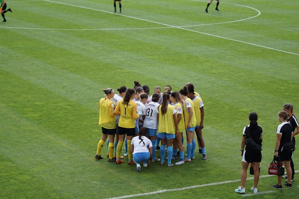 A group of female soccer players, wearing yellow and white jerseys, stands in a tight huddle on a lush green soccer field. The players are focused and engaged, suggesting they are discussing strategies or motivating each other. Nearby, two coaches in black sportswear observe and provide guidance.