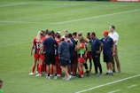 A group of female soccer players and coaches stand together in a huddle on a grassy field. The players are wearing red and black uniforms with some wearing green bibs. The coaches are in blue shirts, and one wears a red cap. The setting appears to be a sports event, possibly before or after a match.