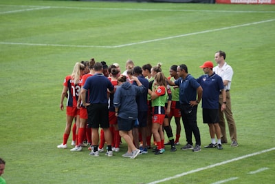 A group of female soccer players and coaches stand together in a huddle on a grassy field. The players are wearing red and black uniforms with some wearing green bibs. The coaches are in blue shirts, and one wears a red cap. The setting appears to be a sports event, possibly before or after a match.