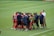 A group of female soccer players and coaches stand together in a huddle on a grassy field. The players are wearing red and black uniforms with some wearing green bibs. The coaches are in blue shirts, and one wears a red cap. The setting appears to be a sports event, possibly before or after a match.
