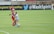 Two soccer players are engaged in a close match on the field, with one player in a blue and white uniform tackling another player in a red and black uniform. The soccer ball is visible near their feet, and another player in red is in the background. The field is lush green and marked with white lines. The backdrop includes advertisements from soccer organizations.