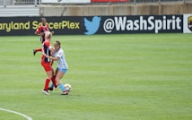 Two soccer players are engaged in a close match on the field, with one player in a blue and white uniform tackling another player in a red and black uniform. The soccer ball is visible near their feet, and another player in red is in the background. The field is lush green and marked with white lines. The backdrop includes advertisements from soccer organizations.