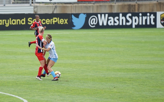 Two soccer players are engaged in a close match on the field, with one player in a blue and white uniform tackling another player in a red and black uniform. The soccer ball is visible near their feet, and another player in red is in the background. The field is lush green and marked with white lines. The backdrop includes advertisements from soccer organizations.
