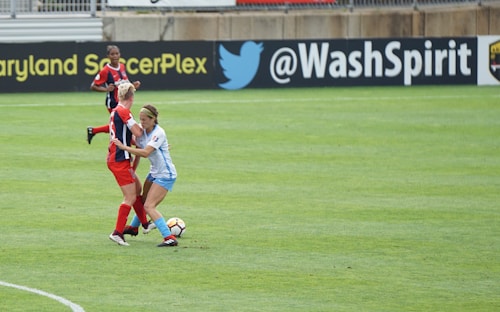 Two soccer players are engaged in a close match on the field, with one player in a blue and white uniform tackling another player in a red and black uniform. The soccer ball is visible near their feet, and another player in red is in the background. The field is lush green and marked with white lines. The backdrop includes advertisements from soccer organizations.