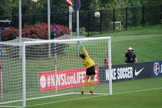 A soccer goalkeeper in a yellow jersey leaps to save a ball heading towards the top of the goal. The scene is set on a grassy pitch with a black fence and some spectators in the background. There is signage on the sidelines, and flags are flying nearby.