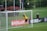 A soccer goalkeeper in a yellow jersey leaps to save a ball heading towards the top of the goal. The scene is set on a grassy pitch with a black fence and some spectators in the background. There is signage on the sidelines, and flags are flying nearby.