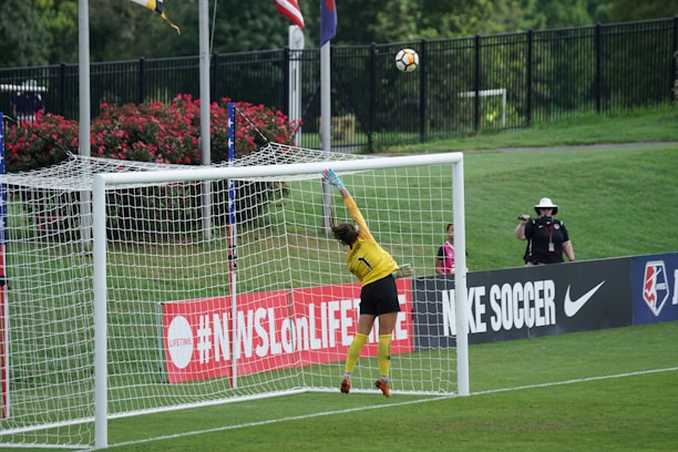 A soccer goalkeeper in a yellow jersey leaps to save a ball heading towards the top of the goal. The scene is set on a grassy pitch with a black fence and some spectators in the background. There is signage on the sidelines, and flags are flying nearby.