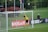 A soccer goalkeeper in a yellow jersey leaps to save a ball heading towards the top of the goal. The scene is set on a grassy pitch with a black fence and some spectators in the background. There is signage on the sidelines, and flags are flying nearby.