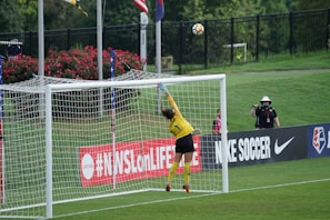 A soccer goalkeeper in a yellow jersey leaps to save a ball heading towards the top of the goal. The scene is set on a grassy pitch with a black fence and some spectators in the background. There is signage on the sidelines, and flags are flying nearby.