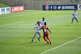 Several female soccer players are actively engaged in a match on a well-maintained grass field. Players are in mid-action, with visible focus and determination. One player in red and blue, labeled with the number 36 and the name McCarty, is interacting with others wearing light blue uniforms. Advertising banners for soccer leagues are displayed in the background along a barrier.