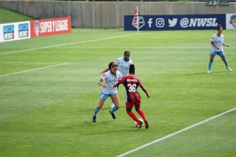 Several female soccer players are actively engaged in a match on a well-maintained grass field. Players are in mid-action, with visible focus and determination. One player in red and blue, labeled with the number 36 and the name McCarty, is interacting with others wearing light blue uniforms. Advertising banners for soccer leagues are displayed in the background along a barrier.