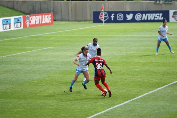Several female soccer players are actively engaged in a match on a well-maintained grass field. Players are in mid-action, with visible focus and determination. One player in red and blue, labeled with the number 36 and the name McCarty, is interacting with others wearing light blue uniforms. Advertising banners for soccer leagues are displayed in the background along a barrier.