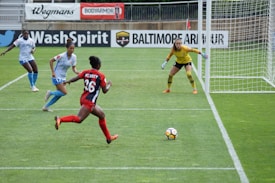 A soccer match in progress with a player in red and black running towards the goal, where a goalkeeper in a yellow jersey is prepared to block the ball. Two other players in white and blue are also visible near the goal.
