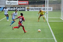 A soccer match in progress with a player in red and black running towards the goal, where a goalkeeper in a yellow jersey is prepared to block the ball. Two other players in white and blue are also visible near the goal.