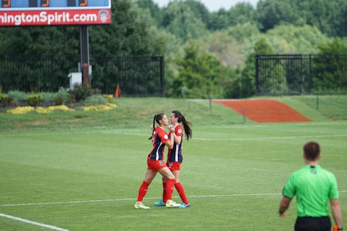 Two soccer players in red and blue uniforms embrace in celebration on a grassy soccer field. A referee in a green jersey stands nearby, and a scoreboard is visible in the background.