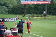 A soccer match scene with a player in a red and blue uniform walking off the field, high-fiving a staff member near the sidelines. A substitution board is held by an official dressed in green. In the background, a scoreboard displays the score, and there is a sign for the Washington Spirit, with greenery and a fence visible further back.
