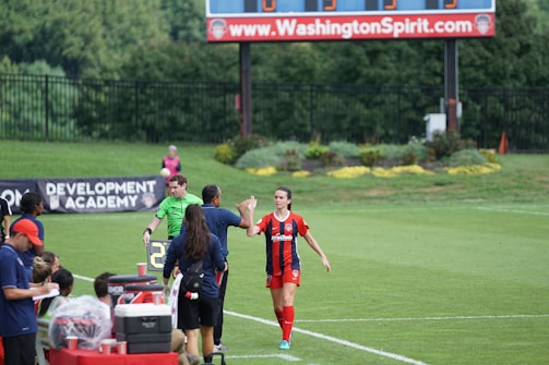 A soccer match scene with a player in a red and blue uniform walking off the field, high-fiving a staff member near the sidelines. A substitution board is held by an official dressed in green. In the background, a scoreboard displays the score, and there is a sign for the Washington Spirit, with greenery and a fence visible further back.