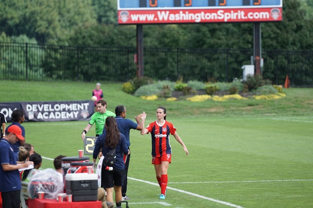 A soccer match scene with a player in a red and blue uniform walking off the field, high-fiving a staff member near the sidelines. A substitution board is held by an official dressed in green. In the background, a scoreboard displays the score, and there is a sign for the Washington Spirit, with greenery and a fence visible further back.