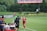 A soccer match scene with a player in a red and blue uniform walking off the field, high-fiving a staff member near the sidelines. A substitution board is held by an official dressed in green. In the background, a scoreboard displays the score, and there is a sign for the Washington Spirit, with greenery and a fence visible further back.