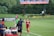A soccer match scene with a player in a red and blue uniform walking off the field, high-fiving a staff member near the sidelines. A substitution board is held by an official dressed in green. In the background, a scoreboard displays the score, and there is a sign for the Washington Spirit, with greenery and a fence visible further back.