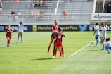 A soccer match in progress, featuring players in red and black uniforms celebrating on the field while others in blue and white observe. The game takes place in a stadium with spectators in the background.