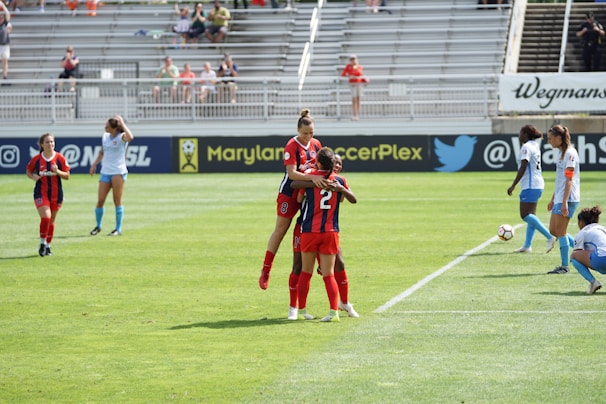 A soccer match in progress, featuring players in red and black uniforms celebrating on the field while others in blue and white observe. The game takes place in a stadium with spectators in the background.