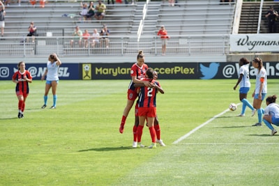 A soccer match in progress, featuring players in red and black uniforms celebrating on the field while others in blue and white observe. The game takes place in a stadium with spectators in the background.
