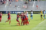 A group of diverse players celebrating a goal on a well-maintained soccer pitch.