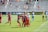 A group of players in red and blue soccer uniforms are celebrating a goal on a soccer field. They are hugging each other with expressions of joy. The background shows a soccer goal, and opposing team members in light blue and a goalkeeper in a yellow uniform. Spectators are visible in the bleachers.