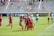 A group of players in red and blue soccer uniforms are celebrating a goal on a soccer field. They are hugging each other with expressions of joy. The background shows a soccer goal, and opposing team members in light blue and a goalkeeper in a yellow uniform. Spectators are visible in the bleachers.