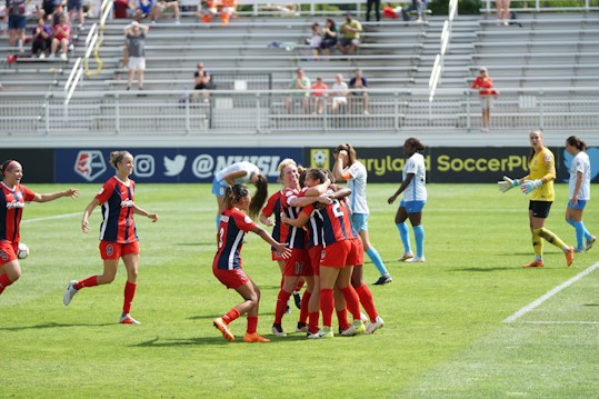 A group of players in red and blue soccer uniforms are celebrating a goal on a soccer field. They are hugging each other with expressions of joy. The background shows a soccer goal, and opposing team members in light blue and a goalkeeper in a yellow uniform. Spectators are visible in the bleachers.