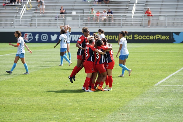 A group of soccer players in red jerseys are celebrating together on a grassy field, while a few players in blue jerseys walk nearby. The setting is an outdoor stadium with bleachers in the background, where spectators are observing the match.