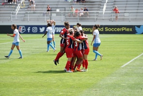 A group of soccer players in red jerseys are celebrating together on a grassy field, while a few players in blue jerseys walk nearby. The setting is an outdoor stadium with bleachers in the background, where spectators are observing the match.