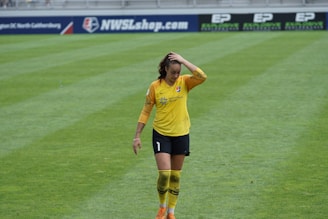 A female soccer player wearing a yellow jersey and black shorts is walking on a grassy field with one hand placed on her head. The background features a sports advertisement board.