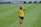 A female soccer player wearing a yellow jersey and black shorts is walking on a grassy field with one hand placed on her head. The background features a sports advertisement board.