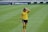 A female soccer player wearing a yellow jersey and black shorts is walking on a grassy field with one hand placed on her head. The background features a sports advertisement board.