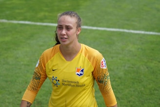 A soccer player wearing a yellow and orange goalkeeper jersey with logos on the front stands on a grassy field. The player has their hair pulled back and appears slightly sweaty, possibly due to physical exertion during a match.