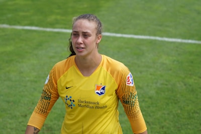 A soccer player wearing a yellow and orange goalkeeper jersey with logos on the front stands on a grassy field. The player has their hair pulled back and appears slightly sweaty, possibly due to physical exertion during a match.