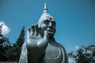 A large stone statue of a serene figure, possibly representing Buddha, prominently displaying an open hand with fingers extended. The statue is set against a clear blue sky, with a few trees visible in the background.