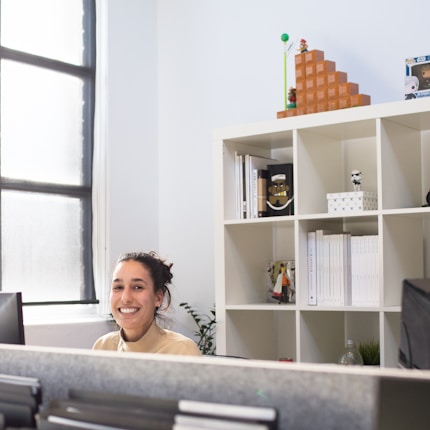 A person is smiling while seated in an office environment. A white shelving unit in the background contains various items, including books, decorative figures, and a small plant. The room is brightly lit with natural light coming through a large window.