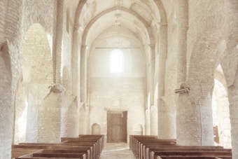 A serene, light-filled interior of a church with high arched ceilings and rows of wooden pews. The walls and ceiling are constructed from light-colored stone bricks, which add a historical and rustic charm to the space. Soft natural light illuminates the area from a window at the back, creating an inviting and peaceful atmosphere.