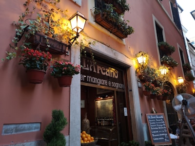 A charming street scene showing the exterior of a cozy restaurant adorned with vibrant flower pots hanging from the walls. Warm, yellow lights illuminate the space, creating a welcoming atmosphere. A menu board is visible on the street, listing several Italian dishes. A small tree is placed near the entrance, adding to the quaint, inviting feel.