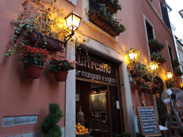A charming street scene showing the exterior of a cozy restaurant adorned with vibrant flower pots hanging from the walls. Warm, yellow lights illuminate the space, creating a welcoming atmosphere. A menu board is visible on the street, listing several Italian dishes. A small tree is placed near the entrance, adding to the quaint, inviting feel.
