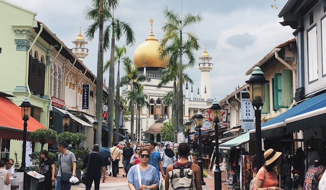 A bustling street scene with a crowd of people walking along a vibrant avenue lined with colorful shopfronts. In the background, a grand mosque with a golden dome and flanking minarets towers above, accented by palm trees reaching into the sky. The atmosphere is lively and filled with diverse individuals engaging with the marketplace.