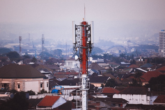 A red and white telecommunications tower stands prominently in the foreground, rising above a densely packed urban area filled with variously colored rooftops and buildings. The skyline extends into a hazy horizon with more buildings and a few additional communication towers visible in the distance.