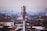 A red and white telecommunications tower stands prominently in the foreground, rising above a densely packed urban area filled with variously colored rooftops and buildings. The skyline extends into a hazy horizon with more buildings and a few additional communication towers visible in the distance.