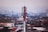 A red and white telecommunications tower stands prominently in the foreground, rising above a densely packed urban area filled with variously colored rooftops and buildings. The skyline extends into a hazy horizon with more buildings and a few additional communication towers visible in the distance.