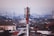 A red and white telecommunications tower stands prominently in the foreground, rising above a densely packed urban area filled with variously colored rooftops and buildings. The skyline extends into a hazy horizon with more buildings and a few additional communication towers visible in the distance.
