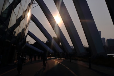 Sunlight streams through large architectural beams, casting long shadows on the pavement below. A reflective glass facade adds to the interplay of light and shadow. People are seen walking and cycling, creating silhouettes against the bright sun.